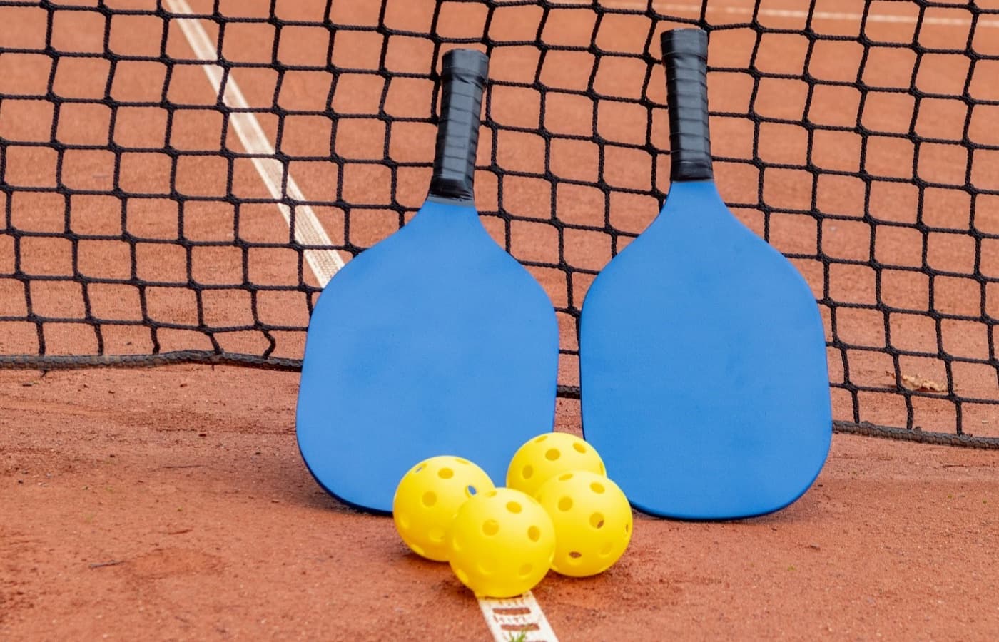 Two pickleball paddles and yellow pickleballs resting by a court net